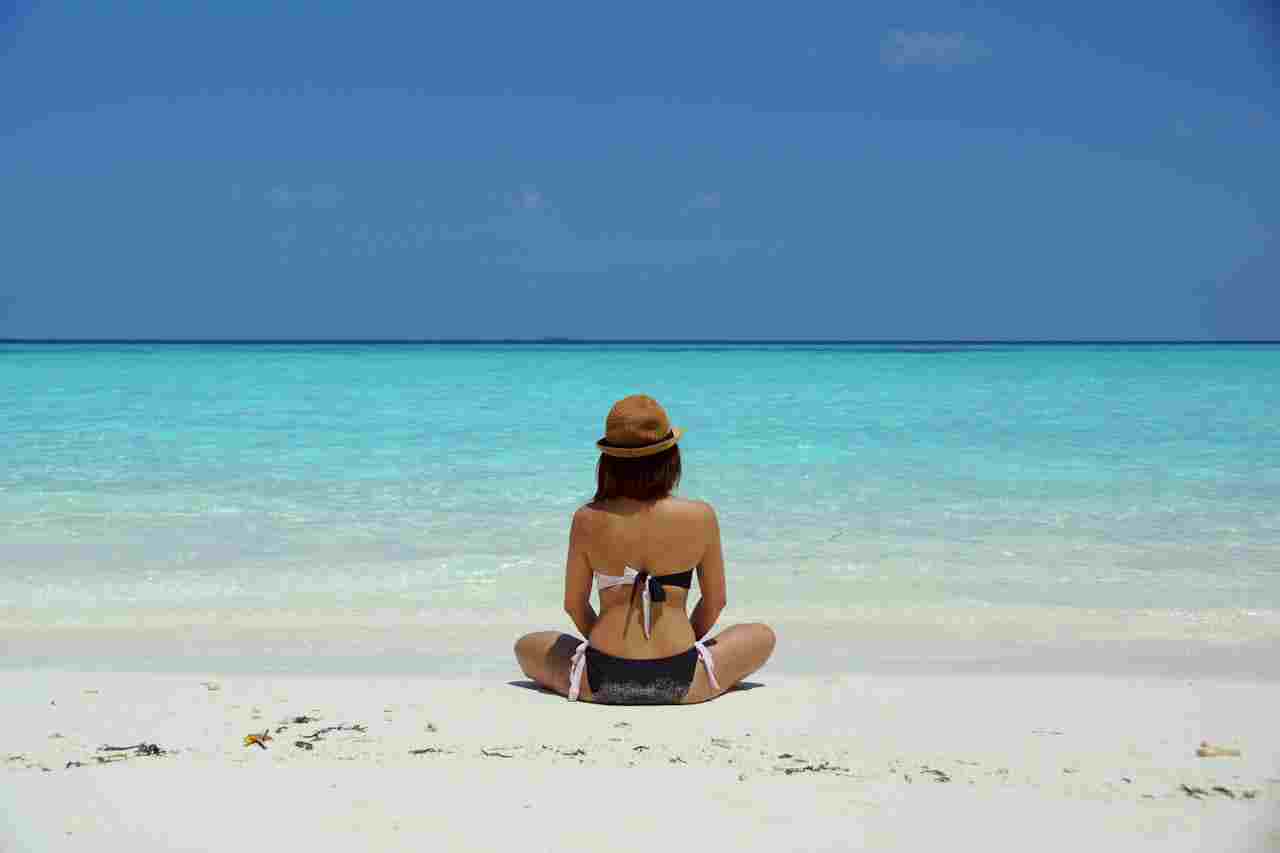 Foto em baixa resolução de uma Mulher sentada em uma praia de areia branca, de costas para a câmera, olhando para o oceano azul-turquesa sob um céu azul claro.